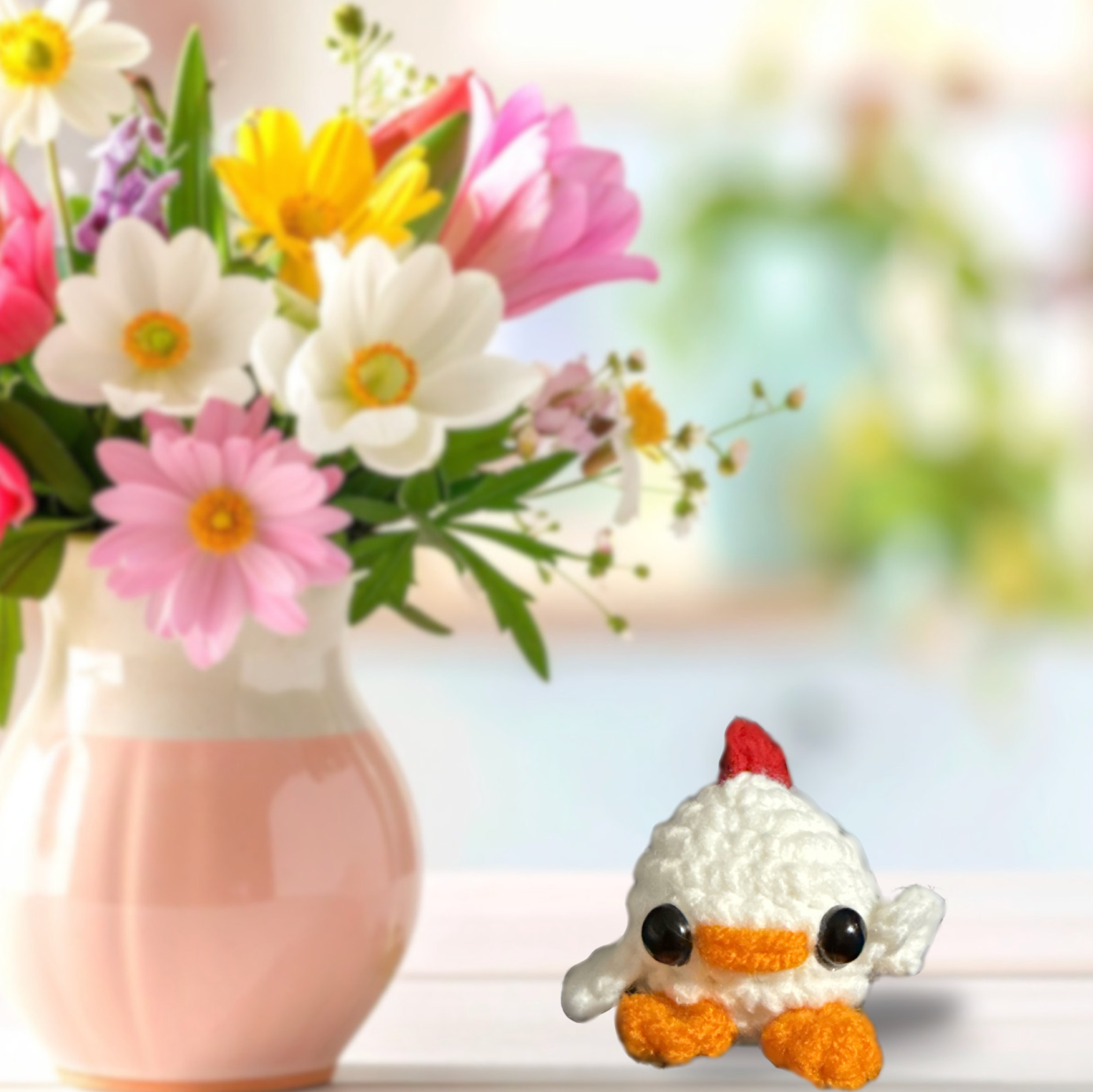 Miracle Nip Chick Cat Toy Lifestyle Image. Small white woven chick cat toy with orange feet, orange beak, red comb, and shiny black eyes displayed on a tabletop beside a blush vase of flowers, showing its decorative, petite scale.