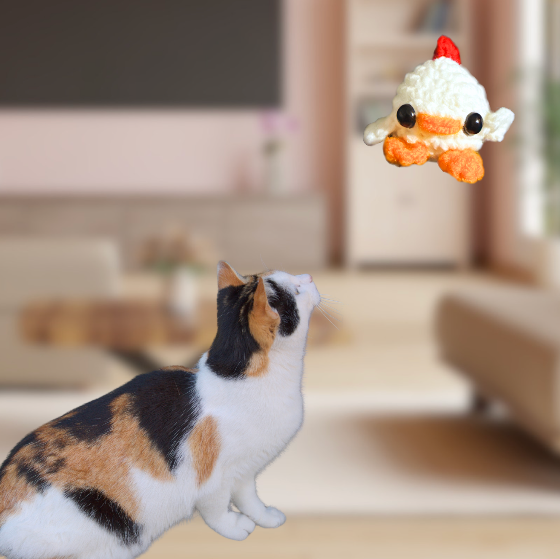 Chick shaped cat Toy with Cat. Small white woven chick cat toy suspended above a calico cat in a softly blurred living room, showing the toy’s tiny scale, rounded shape, and bright orange feet and beak.