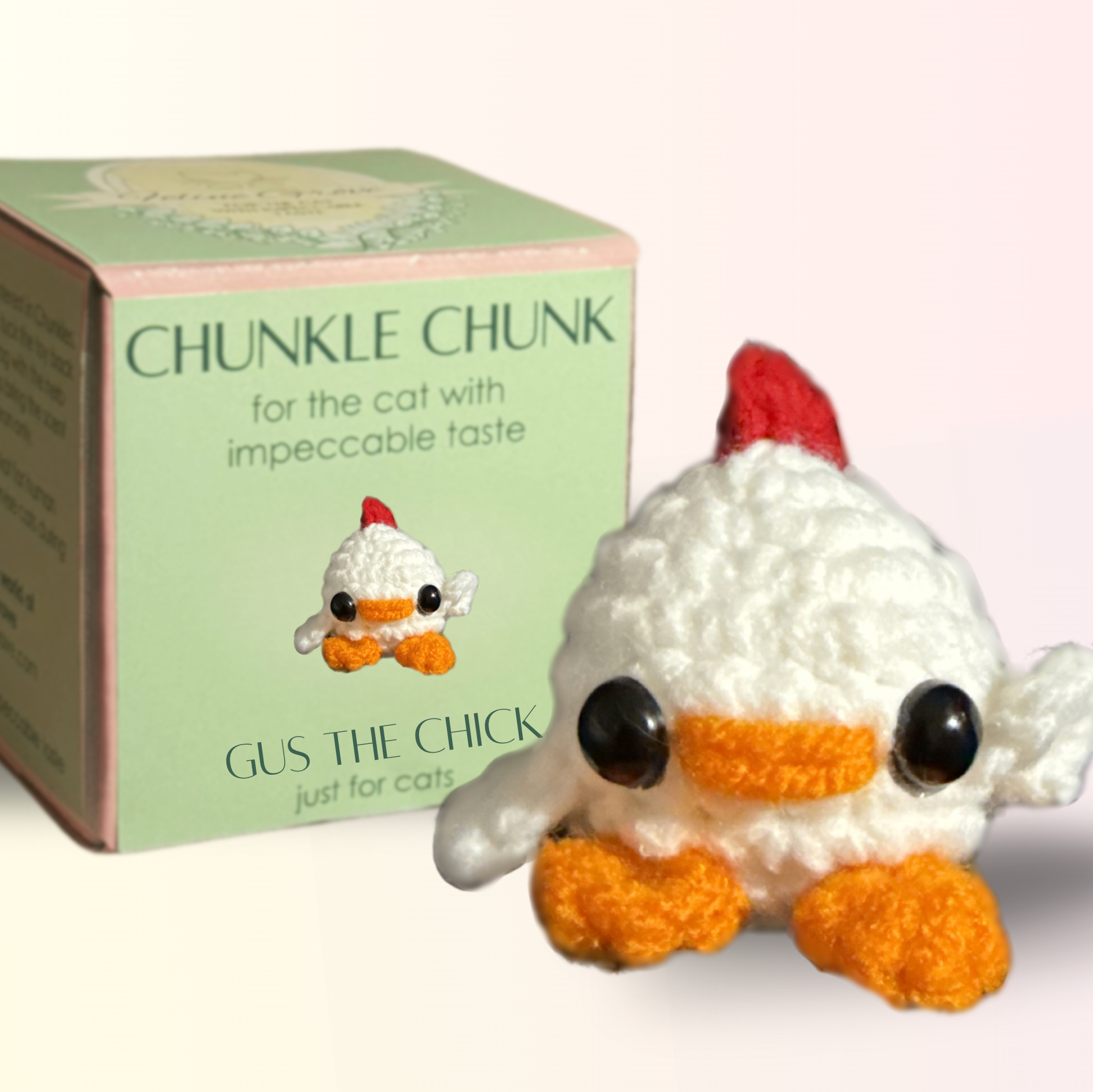 Chick Cat Toy with Packaging. Small white woven chick cat toy with glossy black eyes, orange beak and feet, and a red comb posed beside a pale green Chunkle Chunk box, showing the toy larger than the box illustration.