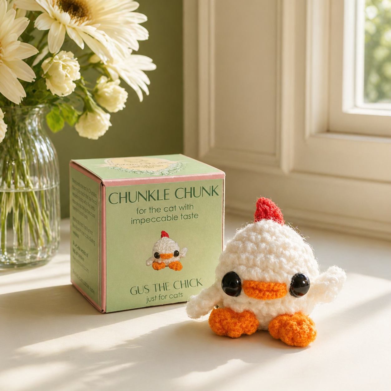 Chick shaped cat toy with packaging. Small white woven chick cat toy with glossy black eyes, orange beak and feet, and a red comb posed beside a pale green Chunkle Chunk box, showing the toy larger than the box illustration.