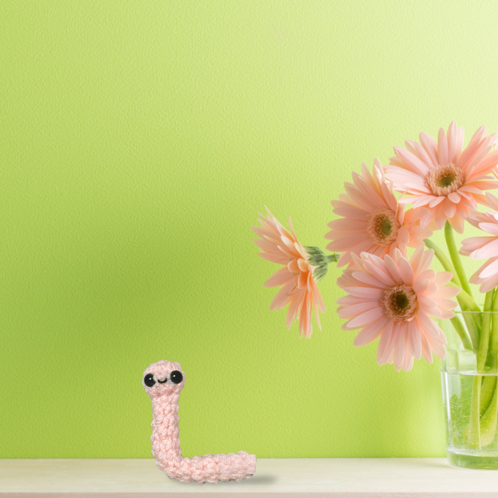Blush pink woven earthworm cat toy displayed on shelf beside soft pink flowers against green background, decorative yet functional Chunkle & Puff luxury cat toy infused with Miracle Nip by Feline Grove