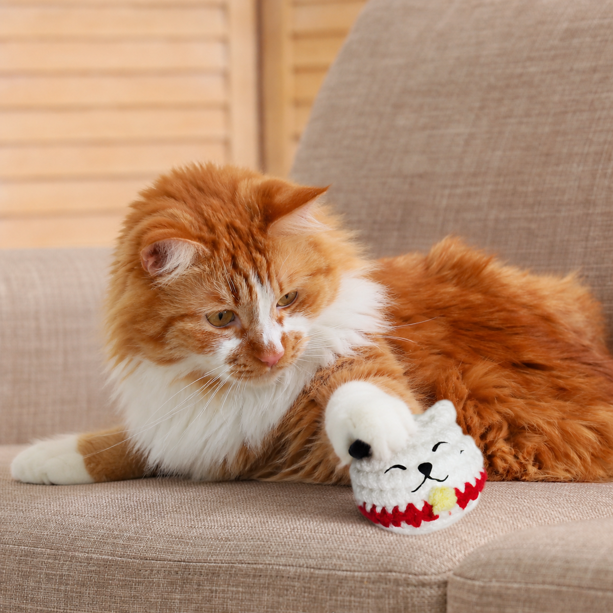 Orange long-haired cat resting on sofa beside small white woven lucky cat toy, plush rounded texture and compact size shown in home setting.