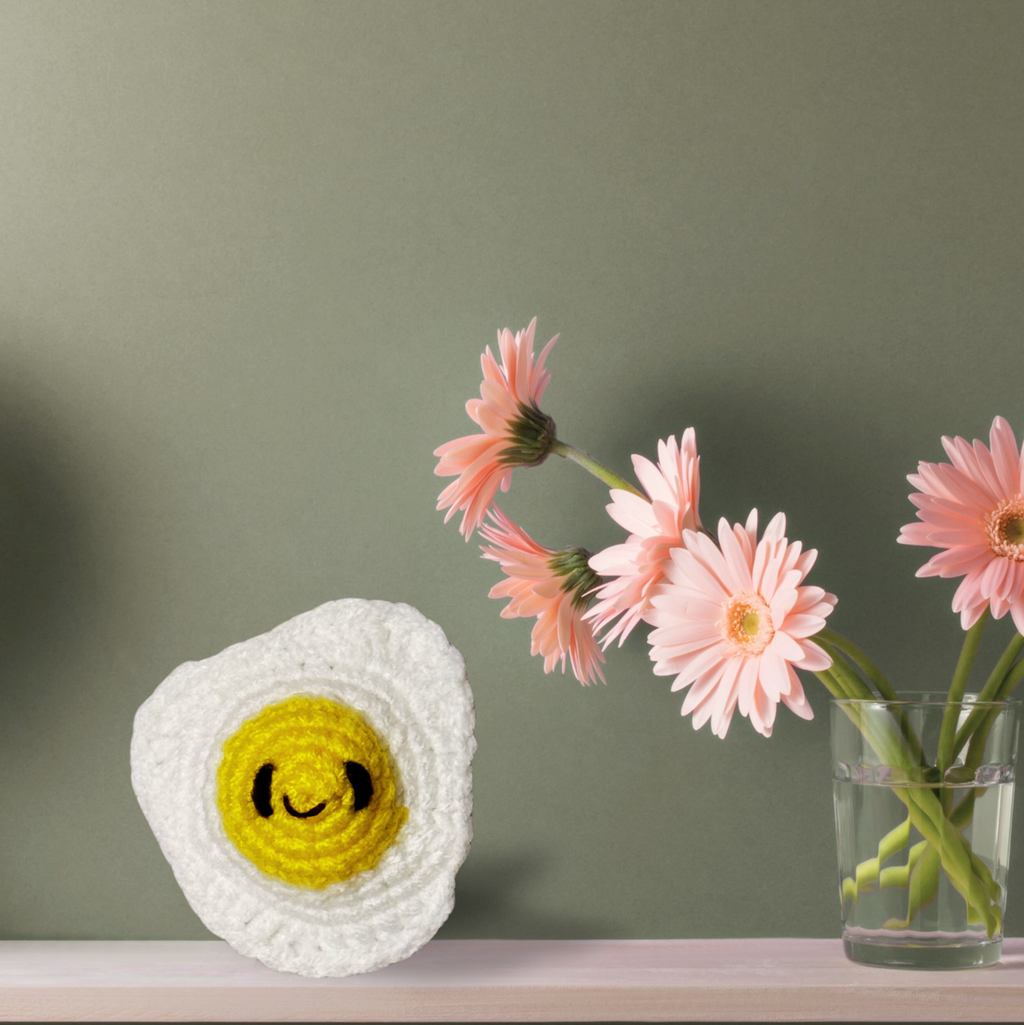 Celeste the Fried Egg Chunkle and Puff cat toy for indoor cats displayed on shelf beside flowers, woven egg shape with soft white texture and yellow center, independent play cat toy with Miracle Nip by Feline Grove including silvervine and valerian