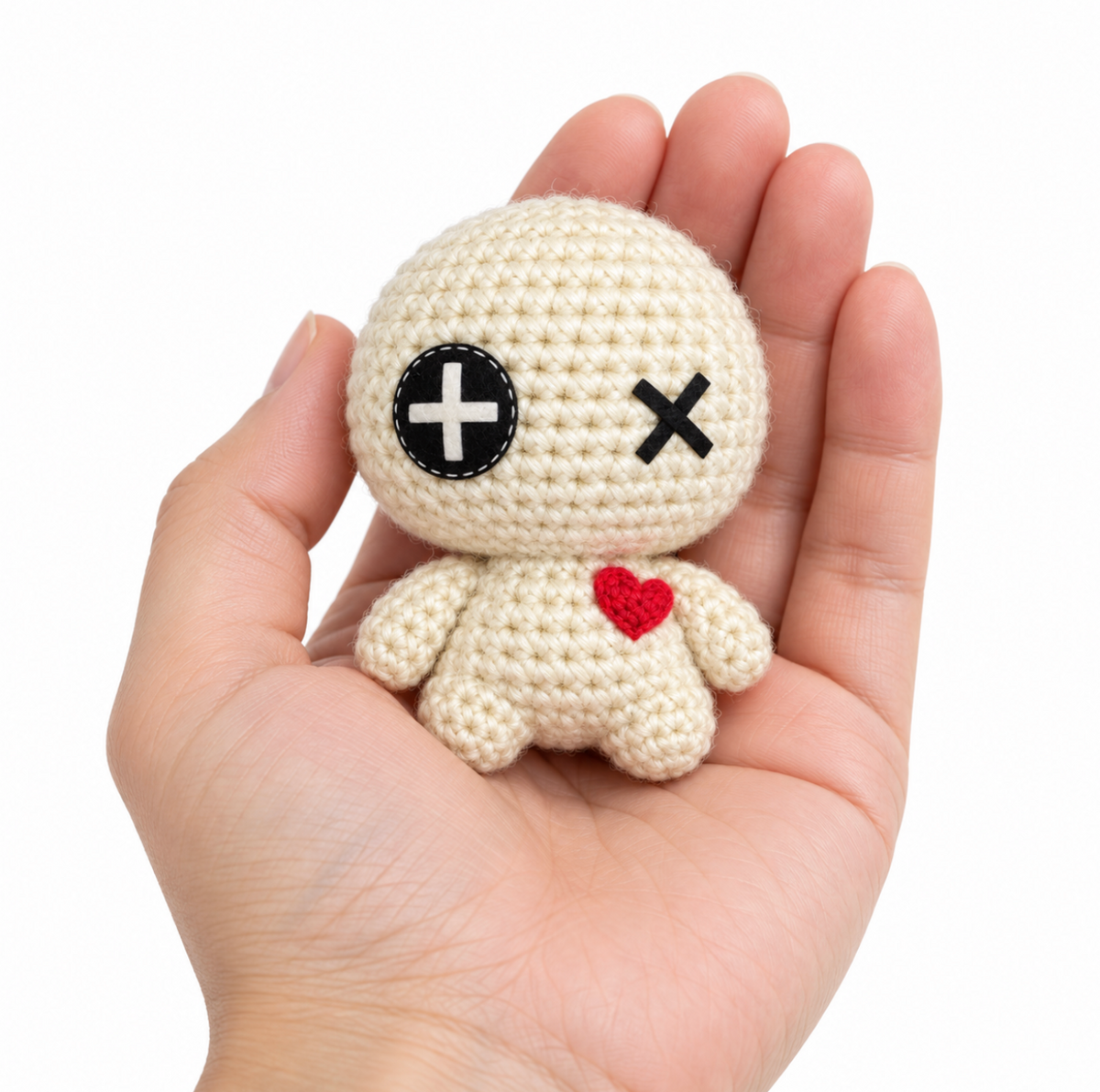 Handmade halloween cat toy voodoo amigurumi doll shown to scale the doll has with a cream body, black stitched eyes, and a red heart on the chest, held in a person's hand against a plain white background.