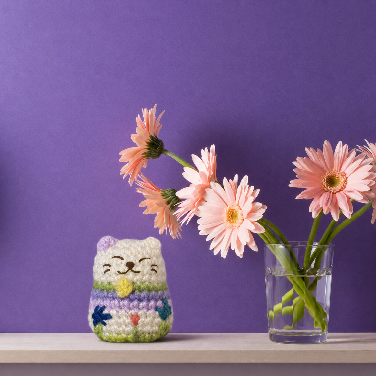 Woven springtime colored lucky cat toy beside glass vase of pink flowers on neutral surface, pastel striped body and embroidered face, compact lightweight design for tossing and display, artisan Chunkle & Puff cat toy infused with Miracle Nip catnip alternative with silvervine