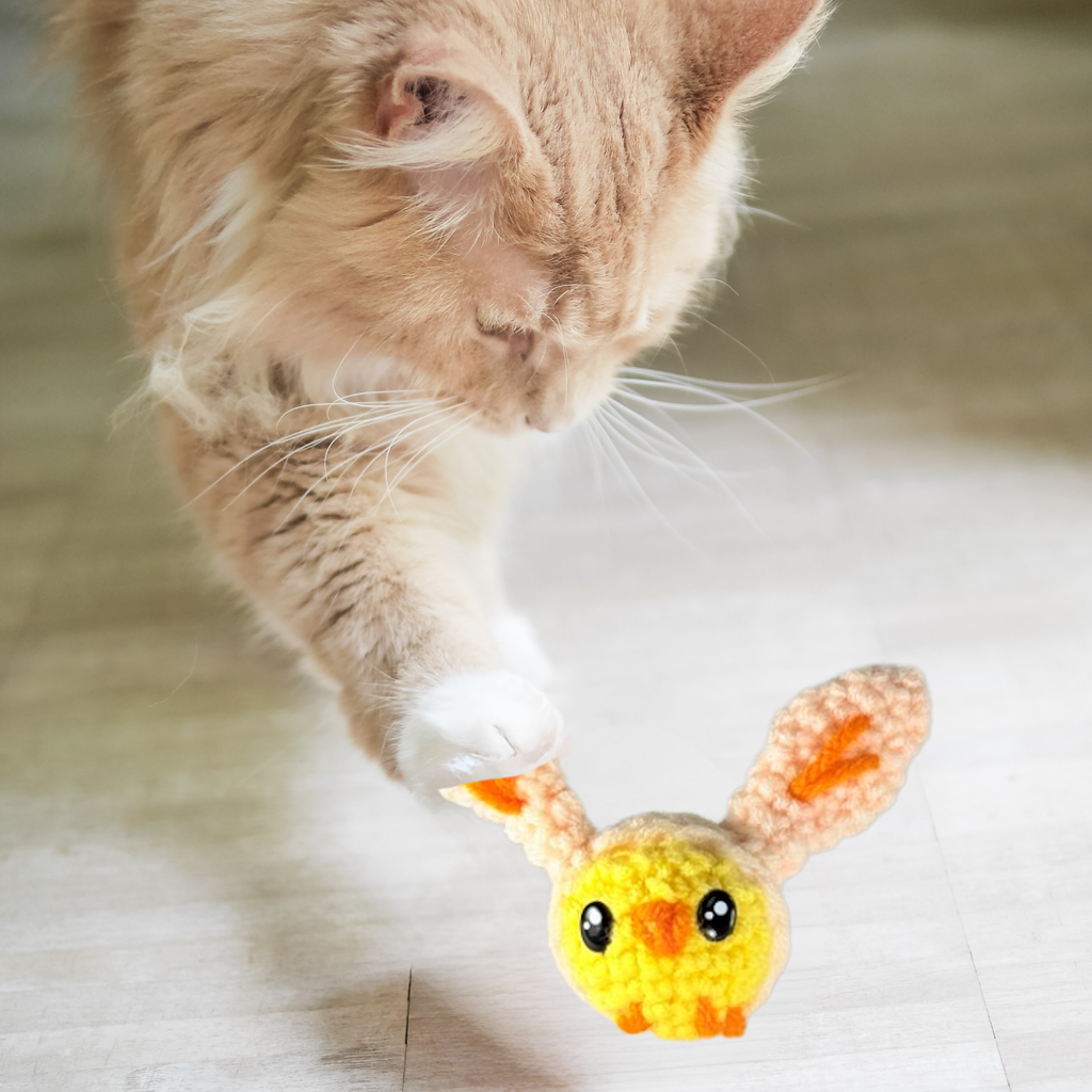 Orange cat reaching out to touch Wally the Chick toy on the floor; soft woven texture, long ears, and small size shown during active engagement.