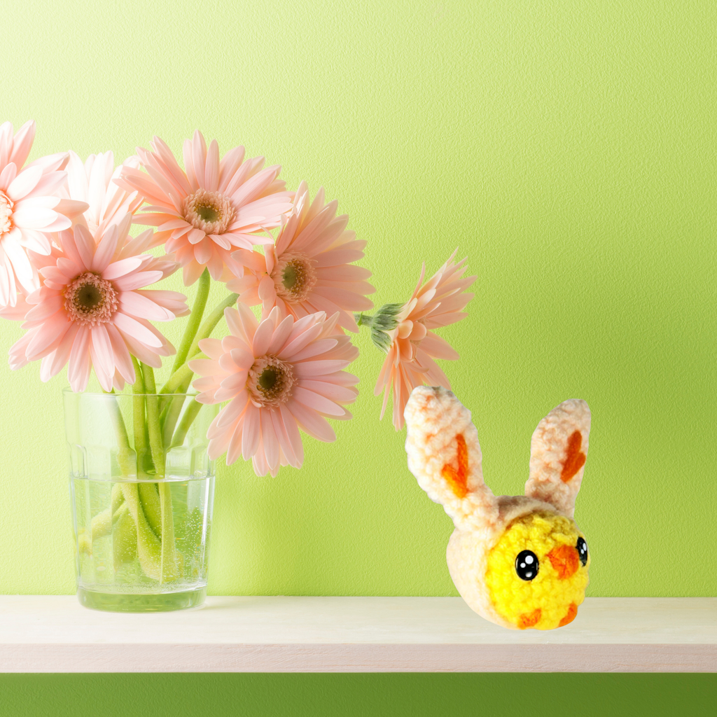 Wally the Chick artisan cat toy displayed on a shelf beside a glass vase of soft pink flowers; small, plush form with textured yarn contrasts with clean, styled home setting.