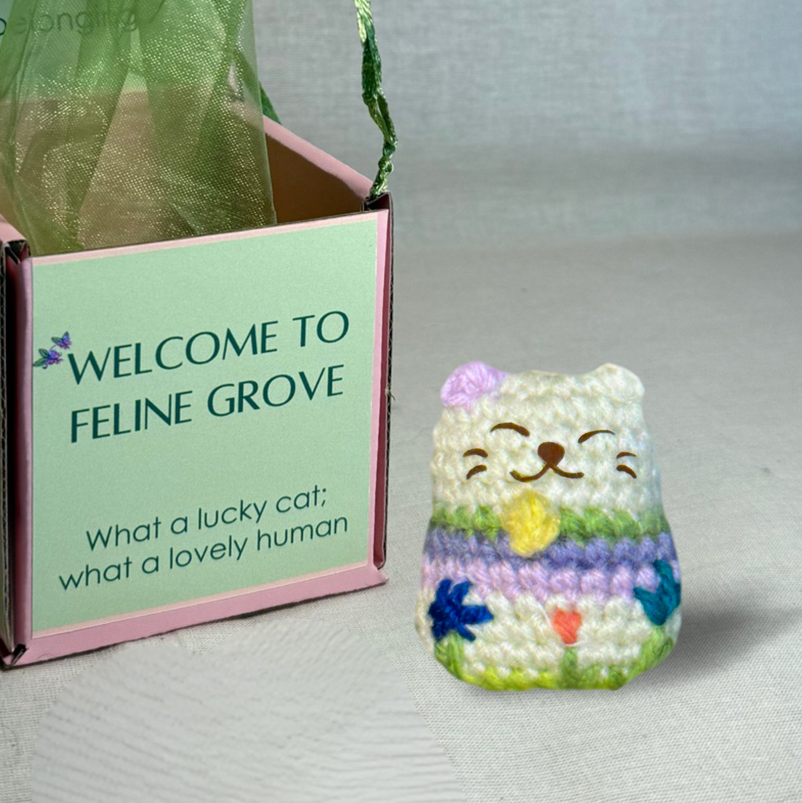 Woven lucky cat toy beside Feline Grove gift box with green organza bag, soft pastel stripes and embroidered face, compact rounded shape for batting and carrying, artisan Chunkle & Puff cat toy infused with Miracle Nip gourmet catnip alternative botanical blend
